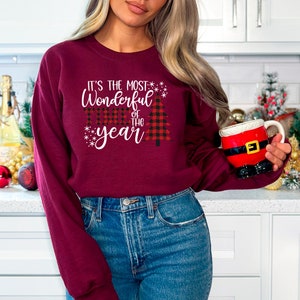 Puede incluir: Una sudadera color burdeos con un gr&aacute;fico blanco que dice "It's the most Wonderful time of the year" con un dise&ntilde;o de &aacute;rbol de Navidad rojo y negro a cuadros.