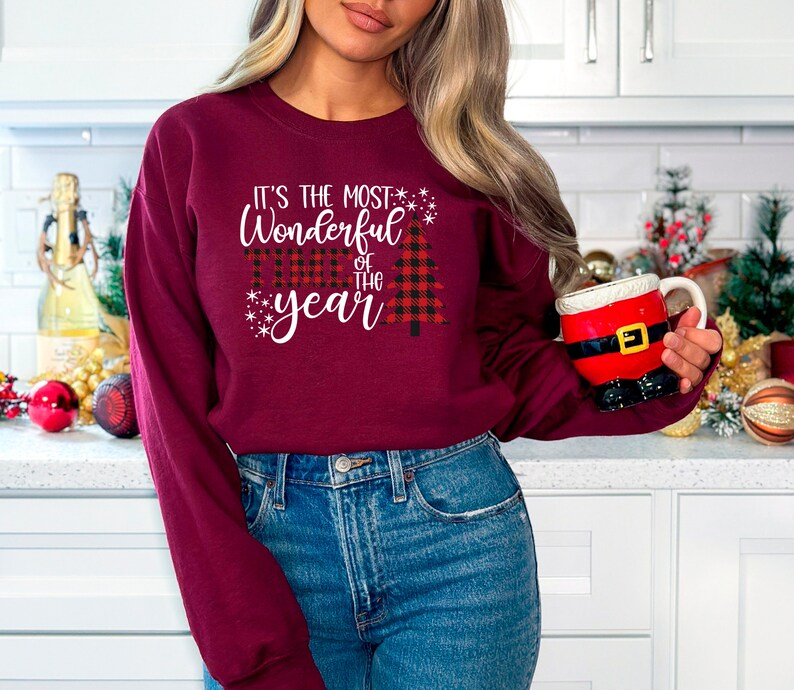 Puede incluir: Una sudadera color burdeos con un gr&aacute;fico blanco que dice "It's the most Wonderful time of the year" con un dise&ntilde;o de &aacute;rbol de Navidad rojo y negro a cuadros.