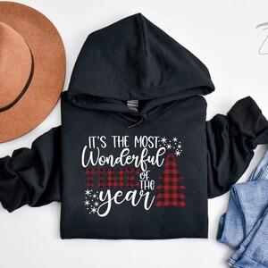 Puede incluir: Sudadera con capucha negra con el texto "It's the most wonderful time of the year" en escritura blanca con un gr&aacute;fico de &aacute;rbol de Navidad de cuadros rojos y negros.