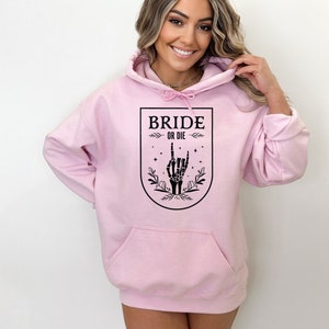 May include: Pink hoodie with a black graphic of a skeleton hand giving the rock and roll sign. The graphic is inside a shield with the text "Bride or Die" above the hand.