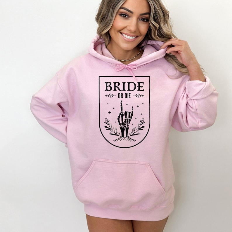 May include: Pink hoodie with a black graphic of a skeleton hand giving the rock and roll sign. The graphic is inside a shield with the text "Bride or Die" above the hand.