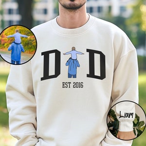 May include: White sweatshirt with a black graphic of the word "DAD" with a small illustration of a father carrying a child on his shoulders. The text "EST 2016" is below the graphic. The sweatshirt has a white cuff with the name "Liam" written in black.
