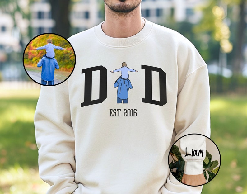 May include: White sweatshirt with a black graphic of the word "DAD" with a small illustration of a father carrying a child on his shoulders. The text "EST 2016" is below the graphic. The sweatshirt has a white cuff with the name "Liam" written in black.