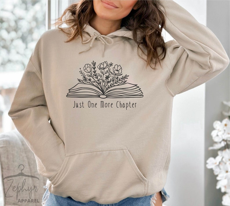 Peut inclure: Un sweat &agrave; capuche beige avec un motif noir d'un livre ouvert avec des fleurs qui en sortent. Le texte "Just One More Chapter" est imprim&eacute; sous le livre.