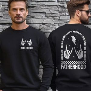 May include: Black sweatshirt with a white graphic print of two skeleton hands making the rock and roll hand gesture. The text "FATHERHOOD" is printed below the hands. The back of the sweatshirt has the text "SOME DAYS I ROCK IT. SOME DAYS IT ROCKS ME. EITHER WAY WE'RE ROCKIN'" printed in a circle around a lightning bolt graphic. The text "FATHERHOOD" is printed below the graphic.