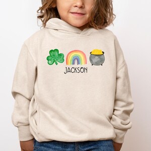 May include: A beige hoodie with a green shamrock, rainbow, and pot of gold design. The text "JACKSON" is printed below the design.