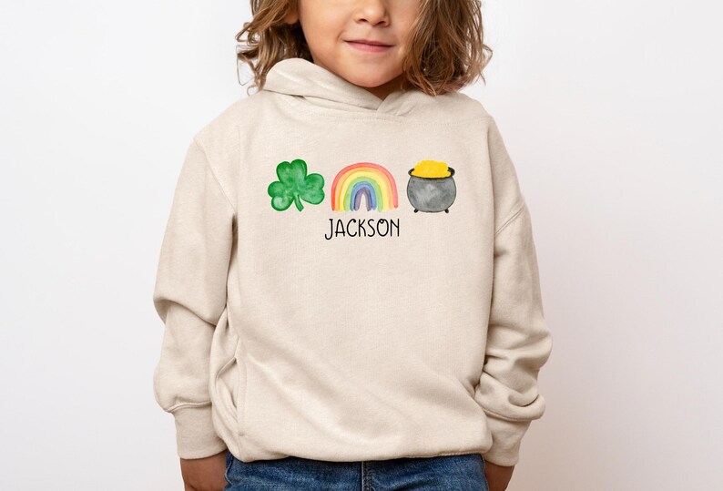 May include: A beige hoodie with a green shamrock, rainbow, and pot of gold design. The text "JACKSON" is printed below the design.