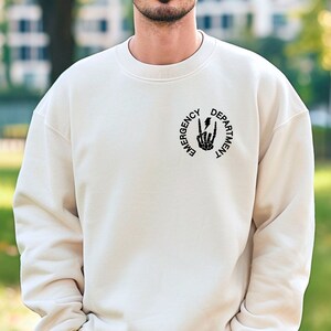 May include: A cream colored sweatshirt with a black graphic of a skeleton hand making a peace sign with a lightning bolt in the middle. The graphic is surrounded by a circle with the text "Emergency Department".