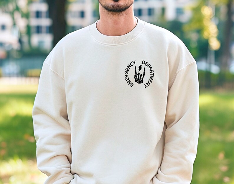May include: A cream colored sweatshirt with a black graphic of a skeleton hand making a peace sign with a lightning bolt in the middle. The graphic is surrounded by a circle with the text "Emergency Department".
