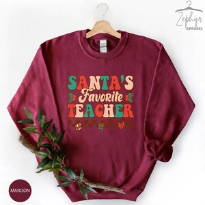 K&ouml;nnte beinhalten: Maroonfarbener Pullover mit dem Text "SANTA'S FAVORITE TEACHER" in einer Retro-Schriftart mit gr&uuml;nen, roten und orangen Farben. Der Text ist von kleinen dekorativen Elementen wie Stechpalmenbl&auml;ttern, Schleifen und Geschenkboxen umgeben.