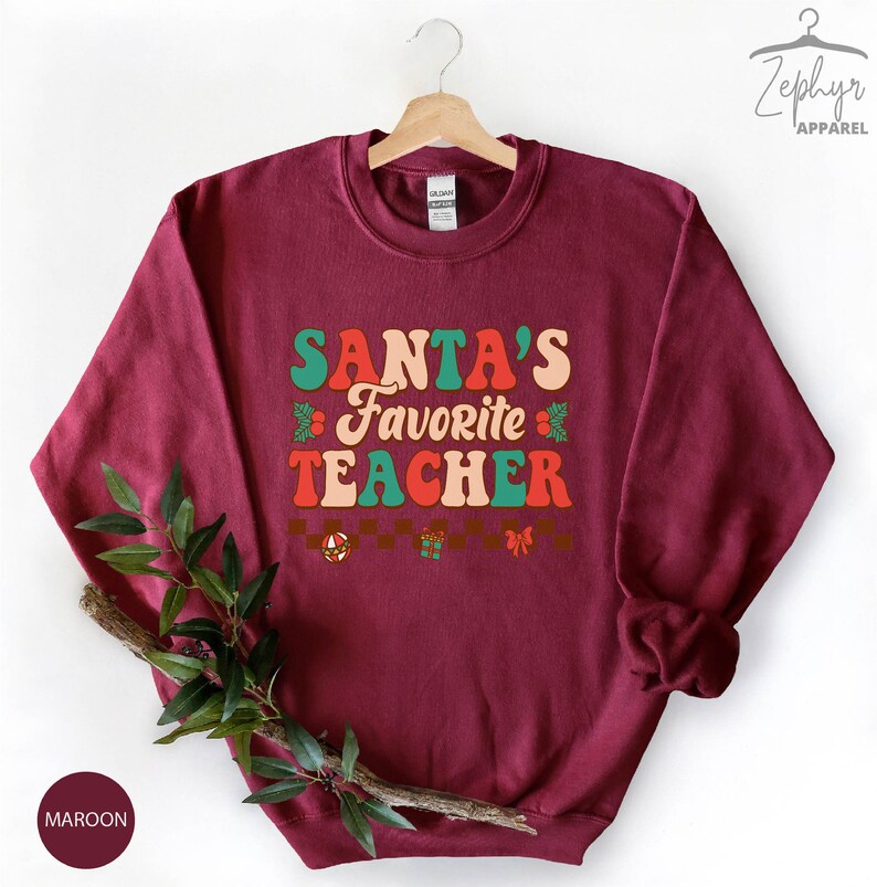 K&ouml;nnte beinhalten: Maroonfarbener Pullover mit dem Text "SANTA'S FAVORITE TEACHER" in einer Retro-Schriftart mit gr&uuml;nen, roten und orangen Farben. Der Text ist von kleinen dekorativen Elementen wie Stechpalmenbl&auml;ttern, Schleifen und Geschenkboxen umgeben.