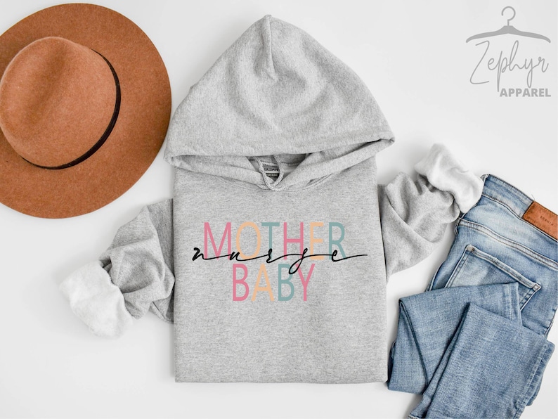 Puede incluir: Una sudadera con capucha gris con el texto "MOTHER nurse BABY" en letras de colores. La sudadera est&aacute; colocada sobre una superficie blanca con un sombrero marr&oacute;n, unos vaqueros azules y una etiqueta blanca con el texto "Zephyr Apparel".