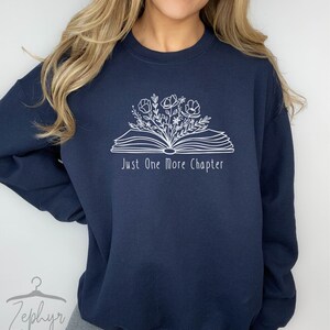 Peut inclure: Un sweat-shirt bleu marine avec un motif blanc d'un livre ouvert avec des fleurs qui en sortent. Le texte "Just One More Chapter" est imprim&eacute; sous le livre.