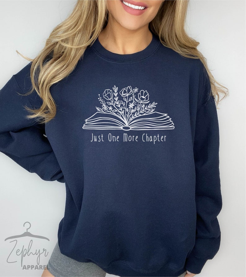 Peut inclure: Un sweat-shirt bleu marine avec un motif blanc d'un livre ouvert avec des fleurs qui en sortent. Le texte "Just One More Chapter" est imprim&eacute; sous le livre.
