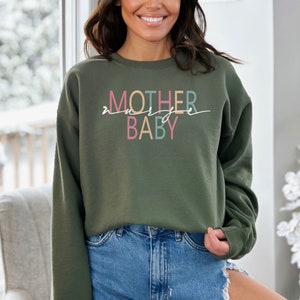 Puede incluir: Una sudadera verde con el texto "MOTHER" en rosa, azul y amarillo, y "BABY" en rosa y azul. El texto est&aacute; escrito en una fuente cursiva.