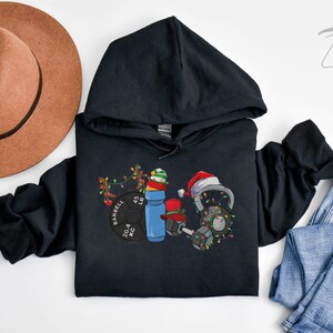 K&ouml;nnte beinhalten: Schwarzer Kapuzenpullover mit einem weihnachtlichen Fitness-Design. Das Design zeigt eine Langhantel, eine Kettlebell, eine Wasserflasche und Weihnachtslichter. Die Langhantel tr&auml;gt den Text "BARBELL 204 KG 45 LB".