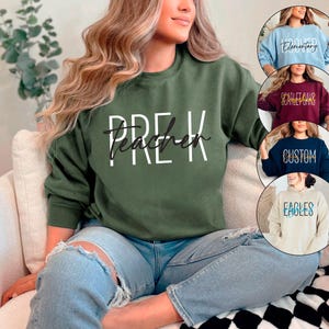 May include: A woman wearing a green sweatshirt with the text "Pre-K Teacher" in white lettering. She is sitting on a white couch with a black and white checkered pattern.