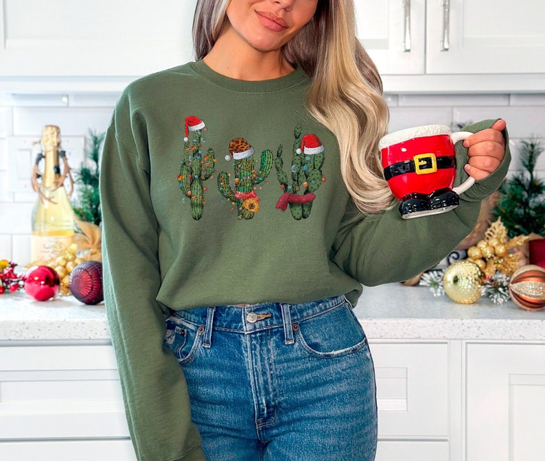 May include: Green Christmas sweatshirt with a graphic of four cacti wearing Santa hats. The cacti are decorated with red and green ornaments. The sweatshirt is being modeled by a person holding a Santa mug.