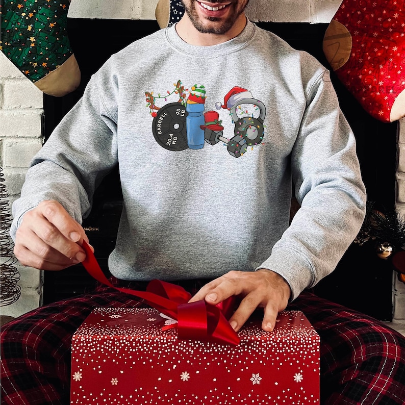 K&ouml;nnte beinhalten: Ein graues Sweatshirt mit einem weihnachtlichen Design, das eine Langhantel, Hanteln, eine Kettlebell, eine Wasserflasche und eine Weihnachtsm&uuml;tze zeigt. Das Design enth&auml;lt Weihnachtslichter und ein Rentier.