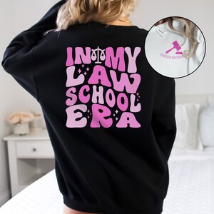 May include: Black sweatshirt with a pink graphic that says "In My Law School Era" with a scale of justice symbol.