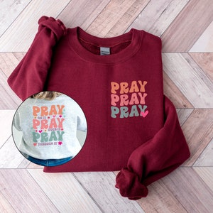May include: A maroon sweatshirt with a white, pink and orange "Pray" graphic on the front. The back of the sweatshirt is grey with a similar graphic that says "Pray on it, Pray over it, Pray through it".