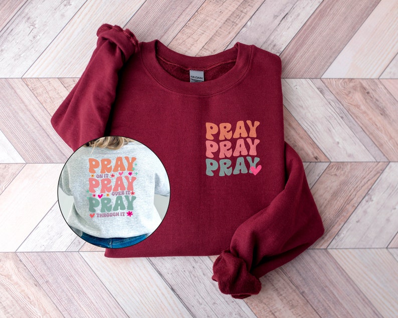 May include: A maroon sweatshirt with a white, pink and orange "Pray" graphic on the front. The back of the sweatshirt is grey with a similar graphic that says "Pray on it, Pray over it, Pray through it".