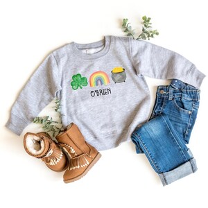 May include: A gray sweatshirt with a green shamrock, rainbow, and pot of gold design. The text "O'Brien" is printed below the design.