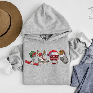 May include: A gray hoodie with a Christmas-themed hockey design. The design features a hockey stick, a hockey skate, a hockey helmet, and a puck, all decorated with Christmas lights and ornaments. The hoodie has a hood and a kangaroo pocket.