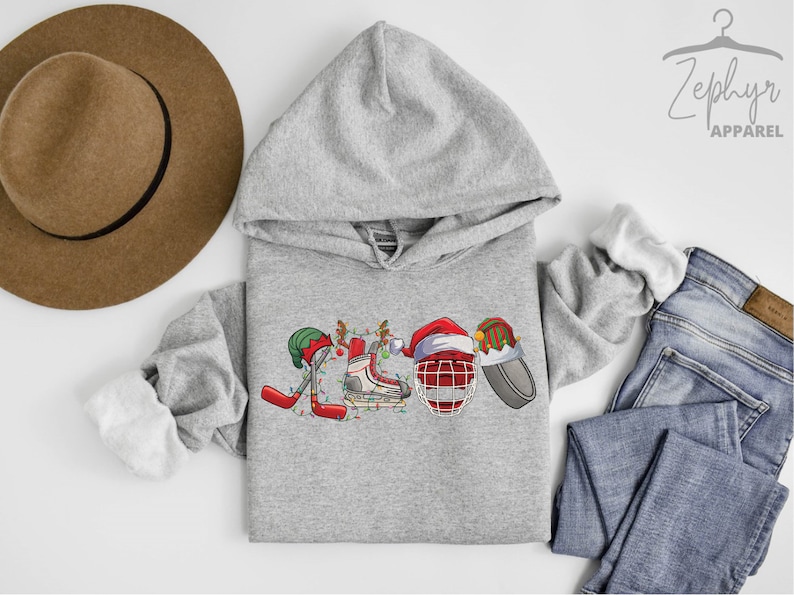 May include: A gray hoodie with a Christmas-themed hockey design. The design features a hockey stick, a hockey skate, a hockey helmet, and a puck, all decorated with Christmas lights and ornaments. The hoodie has a hood and a kangaroo pocket.
