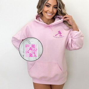 May include: Pink hoodie with a white pocket and a gavel graphic on the front. The text "Custom University" is below the gavel. The back of the hoodie has a pink graphic that says "In My Law School Era".