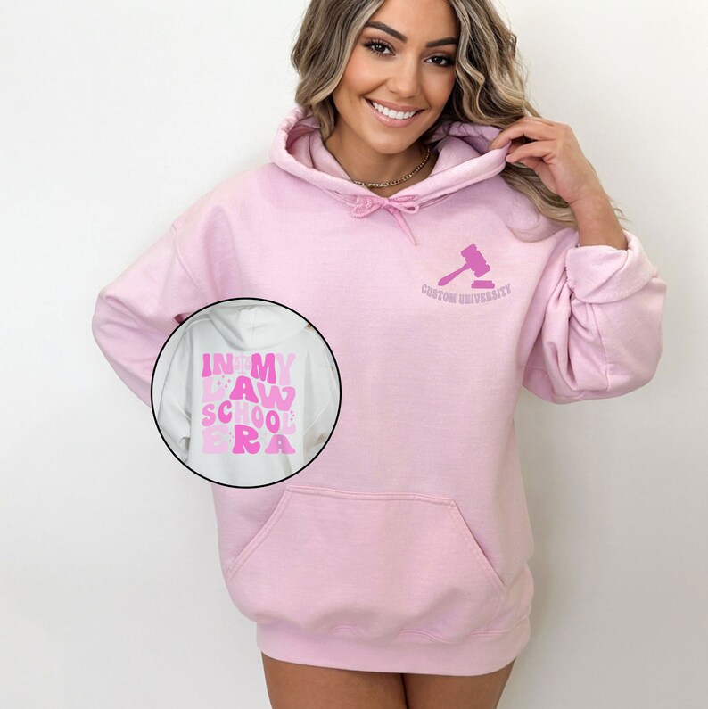 May include: Pink hoodie with a white pocket and a gavel graphic on the front. The text "Custom University" is below the gavel. The back of the hoodie has a pink graphic that says "In My Law School Era".