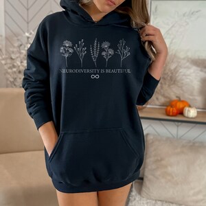 Op de afbeelding: Zwart hoodie met een witte afbeelding van gestileerde bloemen en de tekst "Neurodiversity is Beautiful" met een oneindigheidssymbool.