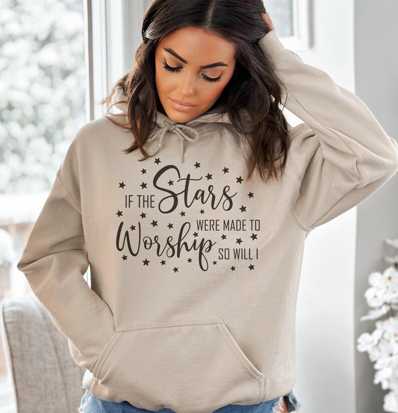 May include: A beige hoodie with a front pocket and drawstring hood. The front of the hoodie features the text "If the Stars Were Made to Worship So Will I" in a black script font, surrounded by small star graphics.