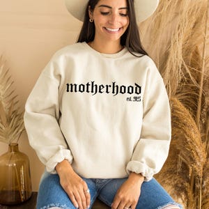 Op de afbeelding: Een cr&egrave;mekleurig sweatshirt met de tekst "motherhood est. 2025" in zwarte gotische letter.