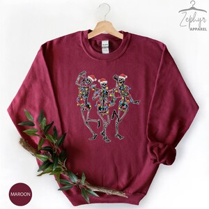 May include: Maroon sweatshirt with a graphic of three skeletons wearing Santa hats and Christmas lights.