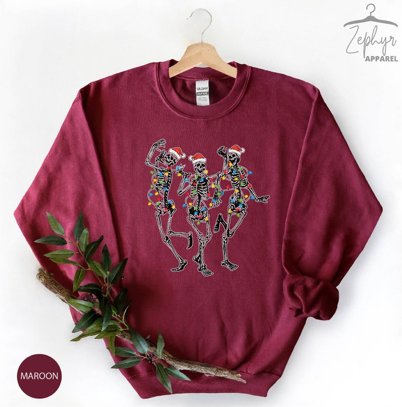 May include: Maroon sweatshirt with a graphic of three skeletons wearing Santa hats and Christmas lights.