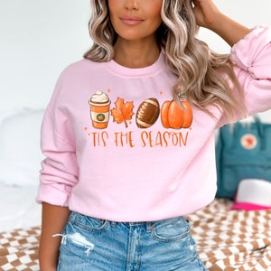 May include: Pink sweatshirt with a graphic of a coffee cup, a leaf, a football, and a pumpkin. The text "'Tis the season" is printed below the graphic.