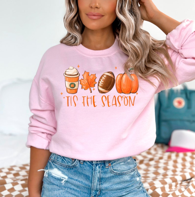 May include: Pink sweatshirt with a graphic of a coffee cup, a leaf, a football, and a pumpkin. The text "'Tis the season" is printed below the graphic.