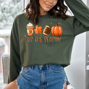 May include: A green sweatshirt with a graphic print of a coffee cup, a leaf, a football, and a pumpkin. The text "Tis the Season" is printed below the graphic.