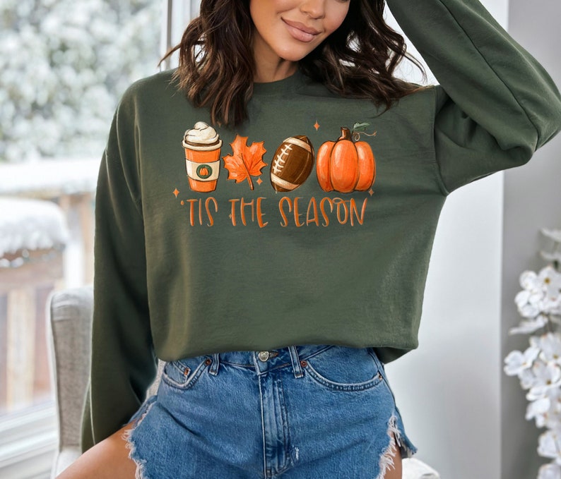 May include: A green sweatshirt with a graphic print of a coffee cup, a leaf, a football, and a pumpkin. The text "Tis the Season" is printed below the graphic.