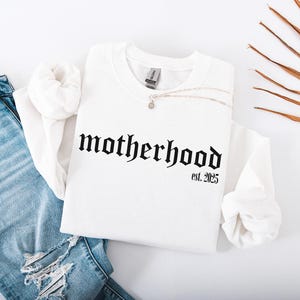 Op de afbeelding: Wit sweatshirt met zwarte gotische letters die "motherhood est. 2025" lezen.