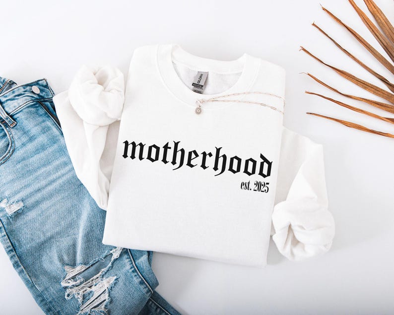 Op de afbeelding: Wit sweatshirt met zwarte gotische letters die "motherhood est. 2025" lezen.