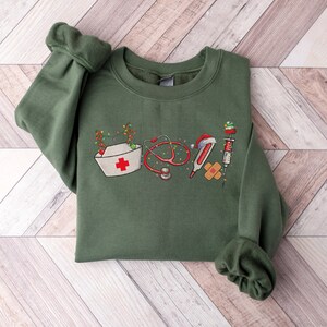 Puede incluir: Sudadera verde con un dise&ntilde;o navide&ntilde;o que incluye herramientas m&eacute;dicas como un estetoscopio, un term&oacute;metro, una venda y una jeringa. El dise&ntilde;o incluye un gorro de enfermera con una cruz roja y una cadena de luces navide&ntilde;as.
