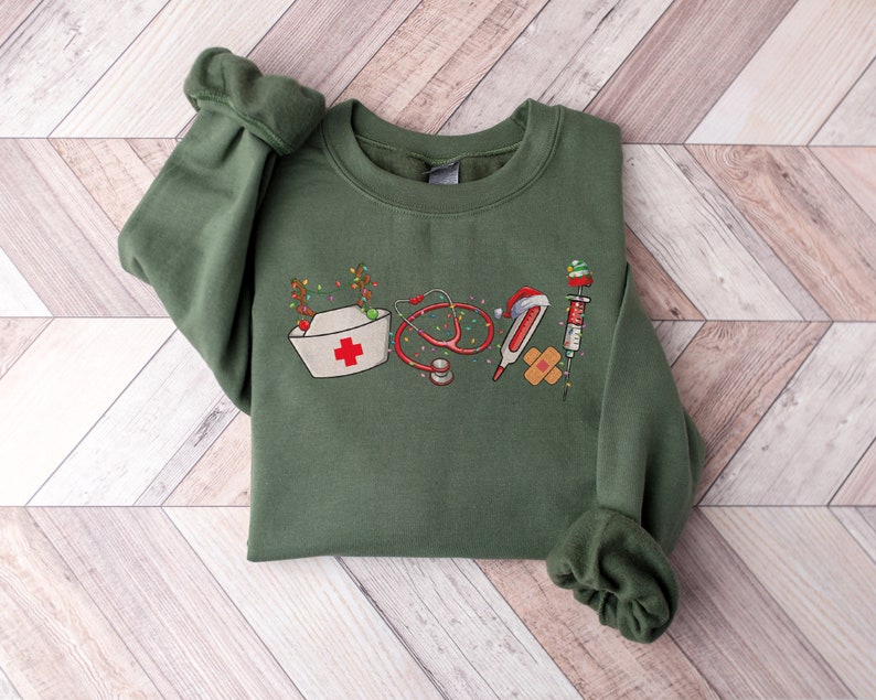 Puede incluir: Sudadera verde con un dise&ntilde;o navide&ntilde;o que incluye herramientas m&eacute;dicas como un estetoscopio, un term&oacute;metro, una venda y una jeringa. El dise&ntilde;o incluye un gorro de enfermera con una cruz roja y una cadena de luces navide&ntilde;as.