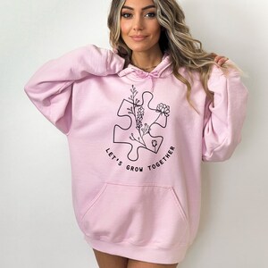 May include: A pink oversized hoodie with a black graphic of a puzzle piece with flowers and the text "Let's Grow Together".