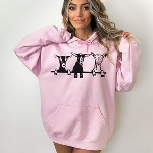 May include: A pink hoodie with a graphic of three goats peeking over a fence. The goats are black and white.