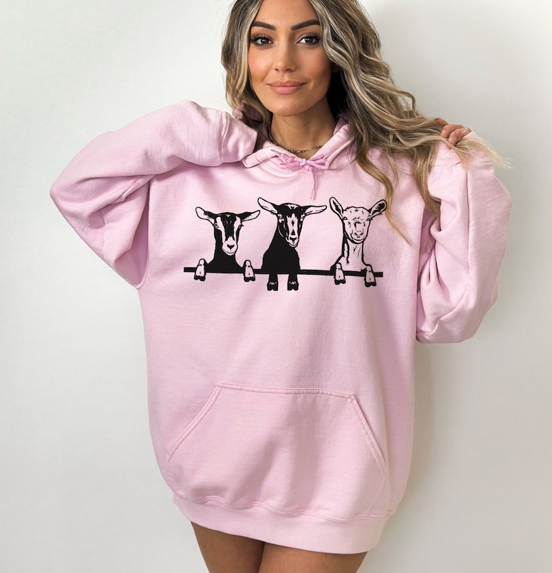 May include: A pink hoodie with a graphic of three goats peeking over a fence. The goats are black and white.