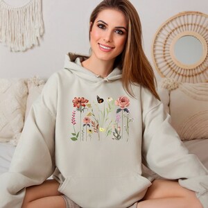 May include: A light beige hoodie with a floral print featuring a butterfly, flowers, and leaves. The hoodie has a drawstring hood and a kangaroo pocket.