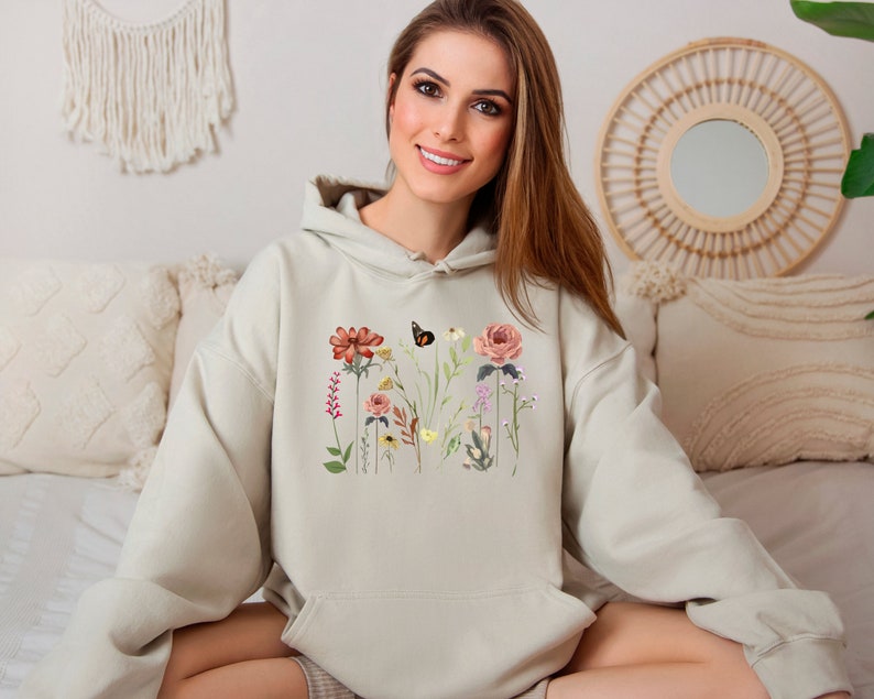 May include: A light beige hoodie with a floral print featuring a butterfly, flowers, and leaves. The hoodie has a drawstring hood and a kangaroo pocket.
