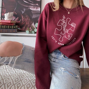 May include: A maroon sweatshirt with a white outline of a puzzle piece with flowers inside. The text "Let's Grow Together" is written around the puzzle piece.